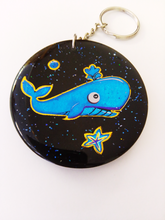 Load image into Gallery viewer, A black, circular keyring or bag ornament made from volcanic ash with a picture of a blue sperm whale, a blue starfish and a blue bubble on a backdrop of blue glitter.